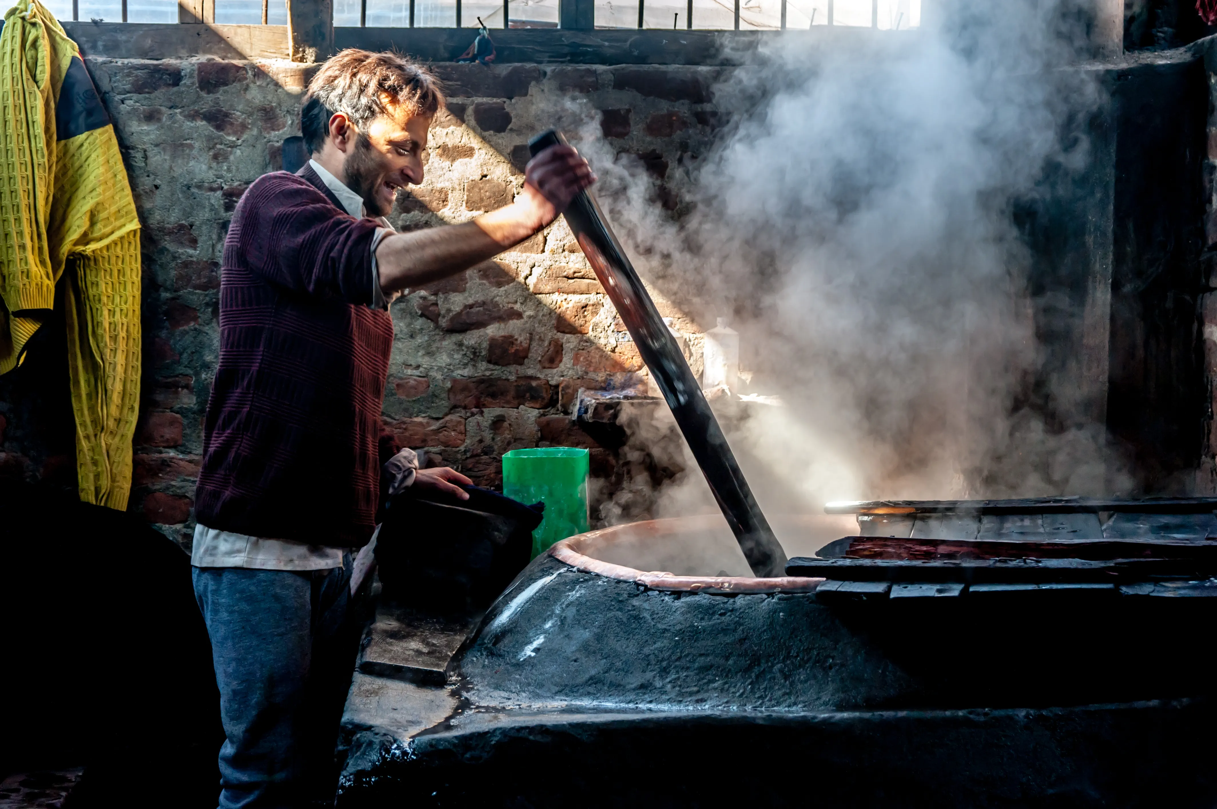 An artisan lifting fabric from a steaming dye vat