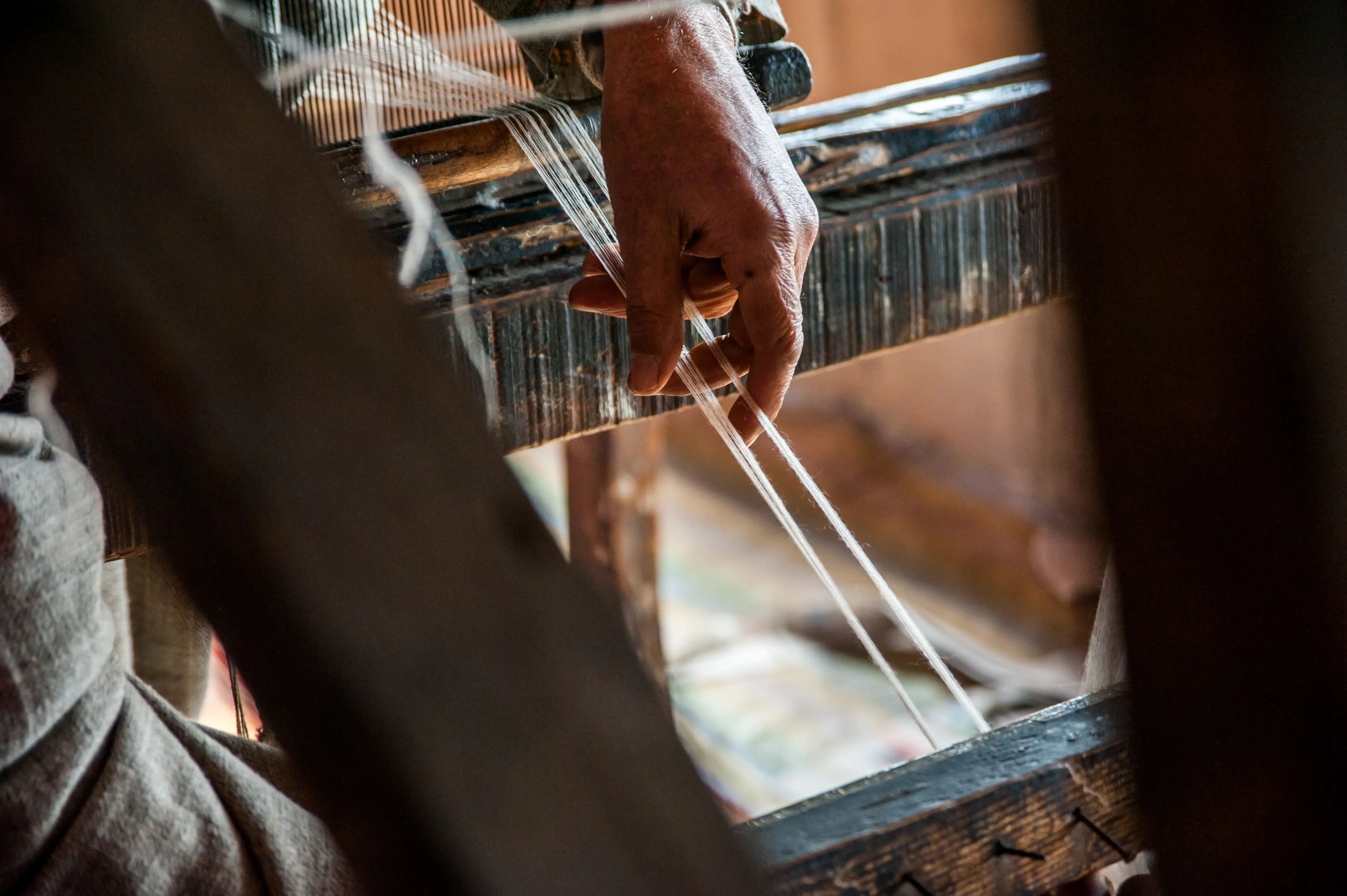 A weaver's hand guiding warp threads through a loom's reed