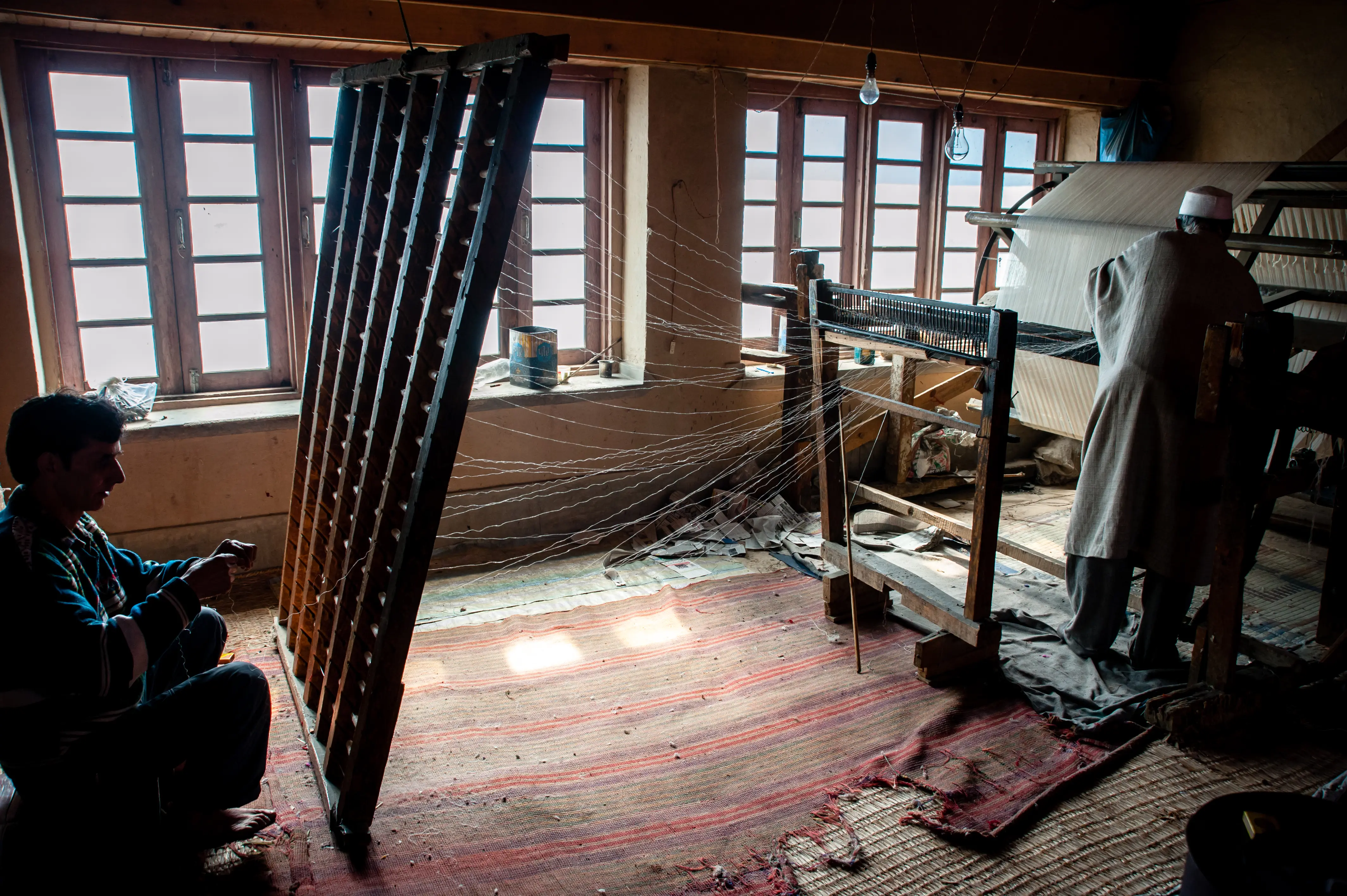 A wide interior view of a Srinagar workshop, a weaver working in silhouette by the window